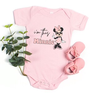 Pu&ograve; includere: Body rosa chiaro per neonati con la scritta "I'm this Minnie" e un disegno di Minnie Mouse. Il disegno mostra Minnie Mouse con occhiali da sole e un vestito a pois. Sono visibili anche scarpine rosa per neonati.
