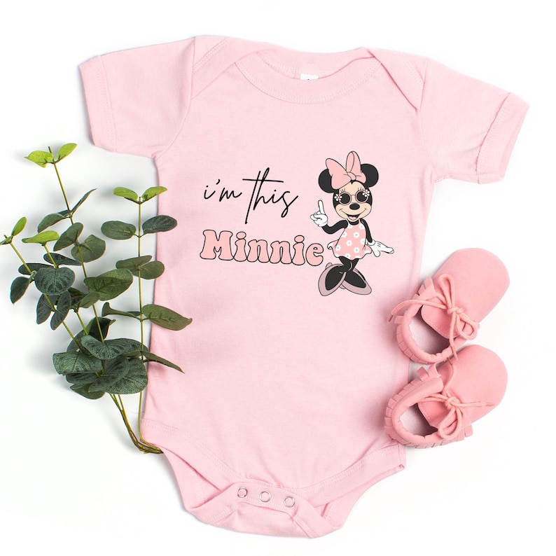 Pu&ograve; includere: Body rosa chiaro per neonati con la scritta "I'm this Minnie" e un disegno di Minnie Mouse. Il disegno mostra Minnie Mouse con occhiali da sole e un vestito a pois. Sono visibili anche scarpine rosa per neonati.