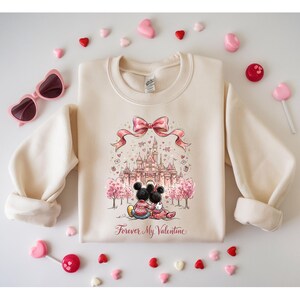 May include: Cream-colored sweatshirt featuring a graphic of Mickey and Minnie Mouse in front of a castle, with the text "Forever My Valentine." The design includes a pink bow and heart accents. Pink heart-shaped sunglasses and candies surround the sweatshirt.