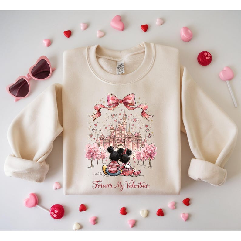 May include: Cream-colored sweatshirt featuring a graphic of Mickey and Minnie Mouse in front of a castle, with the text "Forever My Valentine." The design includes a pink bow and heart accents. Pink heart-shaped sunglasses and candies surround the sweatshirt.