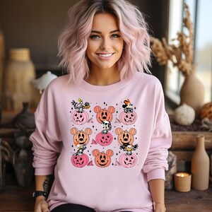 May include: Pink sweatshirt with a Halloween-themed design featuring Mickey Mouse, Minnie Mouse, and Donald Duck dressed as skeletons inside pumpkins.