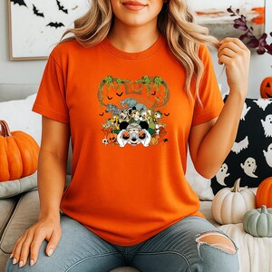 May include: Orange short-sleeved t-shirt featuring a Halloween-themed graphic. The graphic includes a controller, various cartoon animals, and a decorative vine design. The shirt is worn by a person in blue jeans.