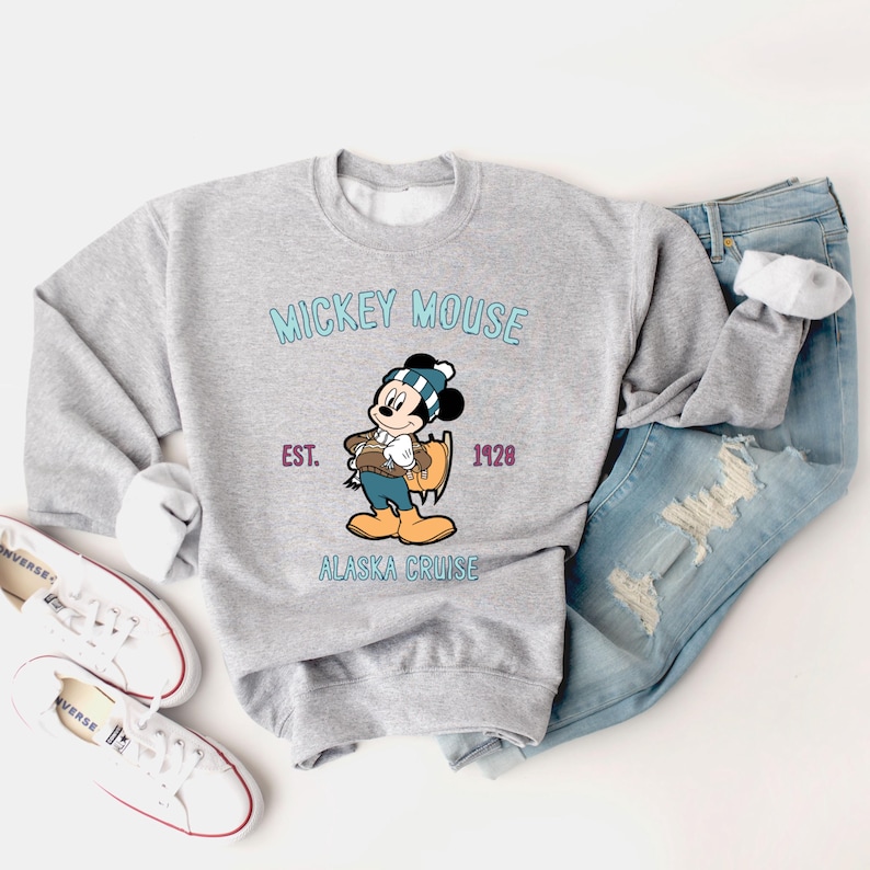 May include: A light gray sweatshirt featuring a graphic of Mickey Mouse in winter attire, with the text "Mickey Mouse Alaska Cruise Est. 1928." The sweatshirt is paired with light blue jeans and white sneakers.