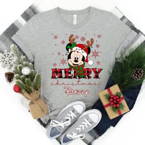 May include: A gray t-shirt with a red and black plaid design. The shirt features a cartoon image of Mickey Mouse wearing a Santa hat and reindeer antlers. The text "Merry Christmas Daddy" is printed on the shirt.