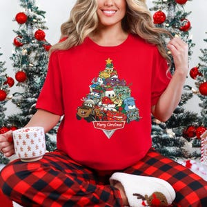 May include: A red short-sleeve t-shirt featuring a Christmas tree design made of cartoon cars. The design includes a gold star on top and the words "Merry Christmas" at the bottom. The person is wearing red and black plaid pajama pants.