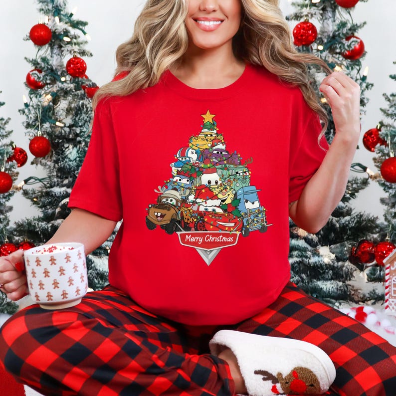 May include: A red short-sleeve t-shirt featuring a Christmas tree design made of cartoon cars. The design includes a gold star on top and the words "Merry Christmas" at the bottom. The person is wearing red and black plaid pajama pants.