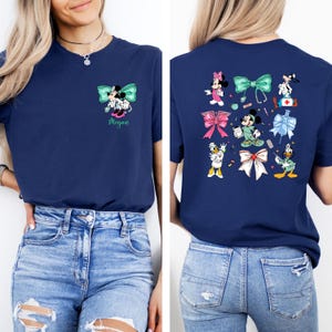 May include: Navy blue t-shirt featuring a Minnie Mouse and Mickey Mouse design with a teal bow and the name "Megan". The back of the shirt displays various cartoon characters and bows. The shirt is made of a soft material.