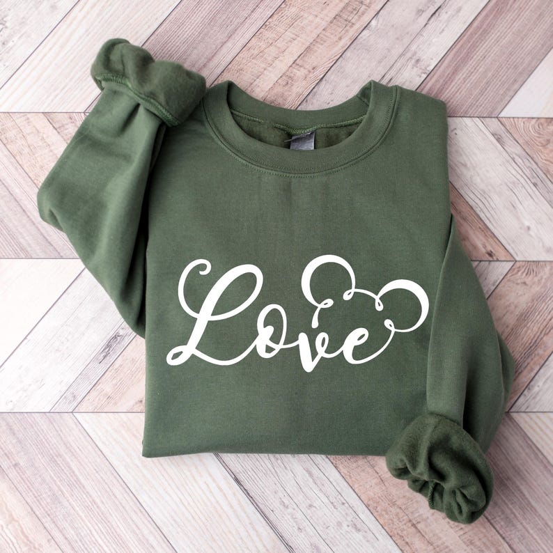 K&ouml;nnte beinhalten: Ein gr&uuml;ner Pullover mit dem Wort "Love" in wei&szlig;er Kursivschrift mit einer Mickey-Maus-Silhouette im "o".