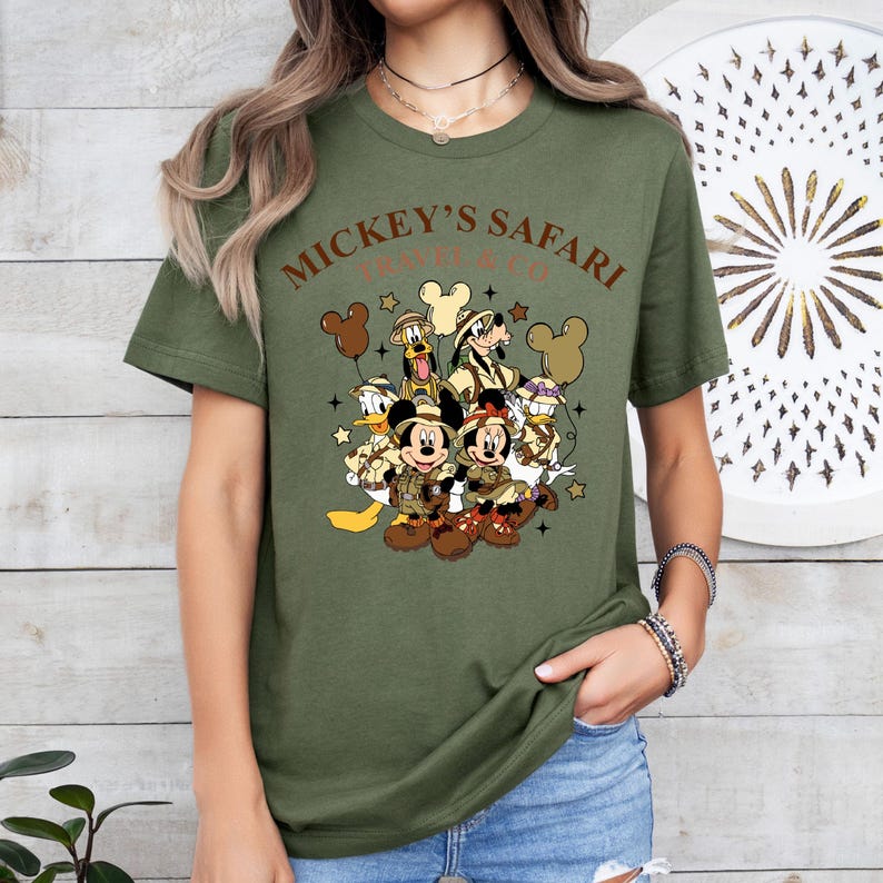 K&ouml;nnte beinhalten: Gr&uuml;nes T-Shirt mit einem grafischen Aufdruck von Mickey Mouse und seinen Freunden in Safari-Outfits. Der Text "Mickey's Safari" ist auf dem Shirt aufgedruckt.
