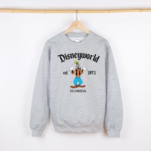 May include: A light gray sweatshirt with the words "Disneyworld" above a cartoon image of Goofy. The image includes the text "est. 1971" and "FLORIDA". The sweatshirt is hanging on a wooden hanger.