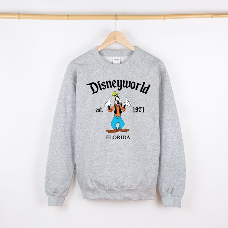 May include: A light gray sweatshirt with the words "Disneyworld" above a cartoon image of Goofy. The image includes the text "est. 1971" and "FLORIDA". The sweatshirt is hanging on a wooden hanger.