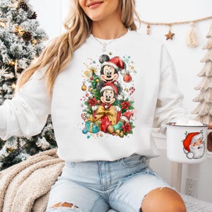 May include: White sweatshirt featuring a festive graphic of Mickey and Minnie Mouse in Santa hats, surrounded by Christmas decorations. The design includes a Christmas tree, presents, and candy canes. A white mug with a Santa Claus illustration is also visible.