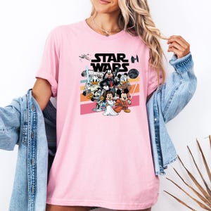 K&ouml;nnte beinhalten: Hellrosa T-Shirt mit einer Grafik von Star Wars-Charakteren, darunter Mickey Mouse, Minnie Mouse, Donald Duck und Goofy. Die Worte "STAR WARS" sind in Schwarz &uuml;ber den Charakteren gedruckt. Das T-Shirt hat kurze &Auml;rmel.