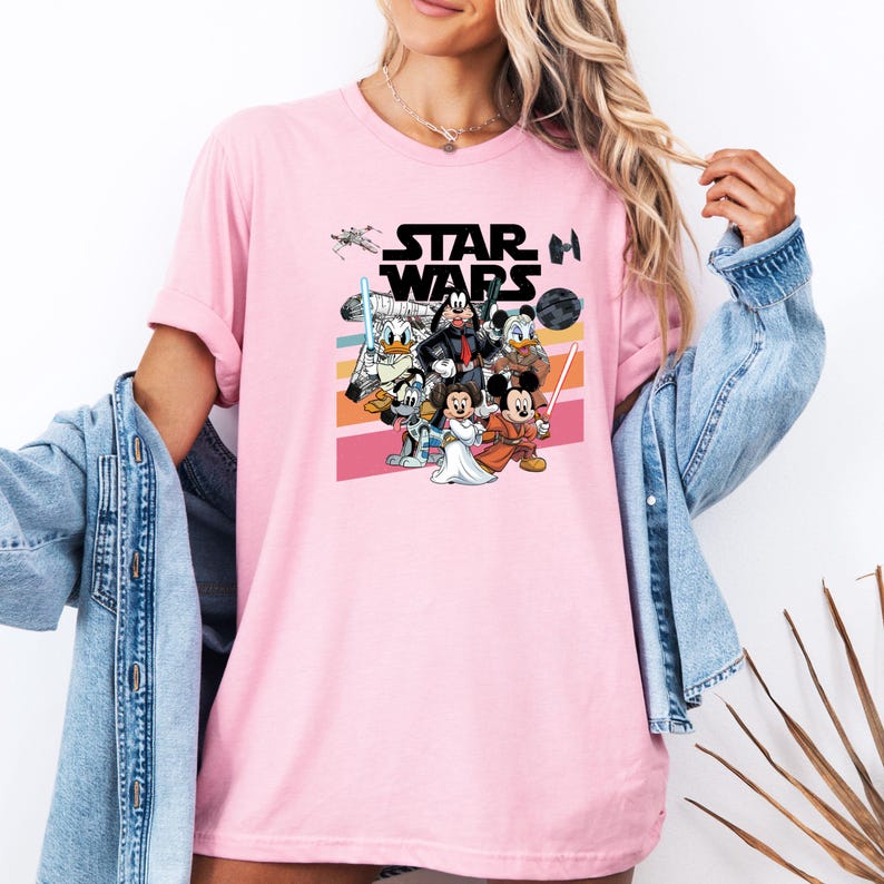 K&ouml;nnte beinhalten: Hellrosa T-Shirt mit einer Grafik von Star Wars-Charakteren, darunter Mickey Mouse, Minnie Mouse, Donald Duck und Goofy. Die Worte "STAR WARS" sind in Schwarz &uuml;ber den Charakteren gedruckt. Das T-Shirt hat kurze &Auml;rmel.
