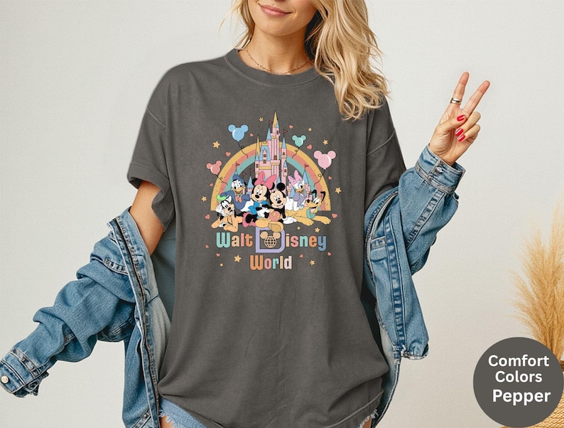 Puede incluir: Camiseta gris oscuro con un gr&aacute;fico colorido de personajes de Disney, un castillo y un arco&iacute;ris. El texto "Walt Disney World" se muestra debajo. La camiseta es de color confort y tiene un corte relajado.