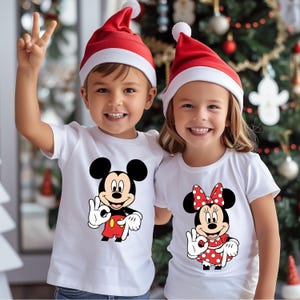 May include: Two children wearing white t-shirts featuring Mickey and Minnie Mouse, and red Santa hats. The boy is giving a peace sign. The girl is smiling and wearing a red polka dot bow. A Christmas tree is in the background.