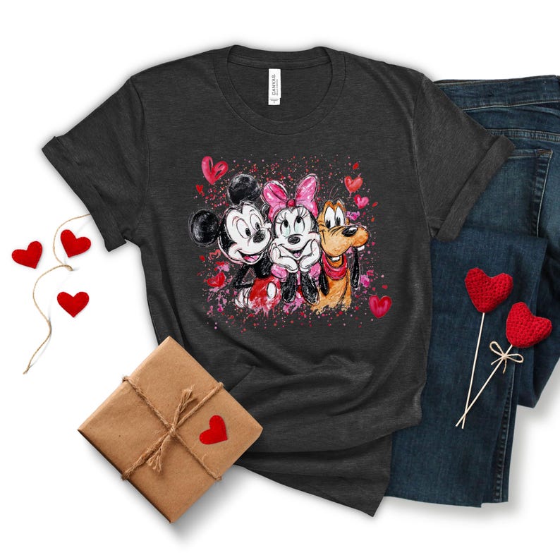 May include: Dark gray t-shirt featuring Mickey Mouse, Minnie Mouse, and Pluto with pink hearts. The graphic design is surrounded by pink paint splatters and hearts. A wrapped gift box and denim jeans are also in the image.