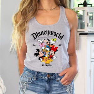 Pu&ograve; includere: Una canotta grigia con una grafica nera di Topolino, Minnie Mouse, Paperino, Pippo e Pluto. La grafica dice "Disneyworld Est 1971 Florida".