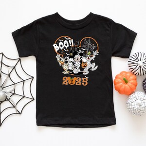 May include: Black short-sleeved t-shirt with a Halloween graphic. The design features Mickey Mouse, Minnie Mouse, and Goofy as mummies, with the text "BOO!!" and "2025" in orange. A spider web and a haunted house are also included in the design.