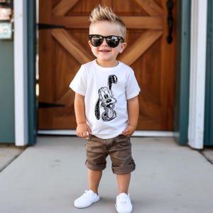 May include: A young child wearing a white t-shirt with a black and white cartoon dog graphic, brown shorts, white sneakers, and black sunglasses. The child is standing outside in front of a wooden door.
