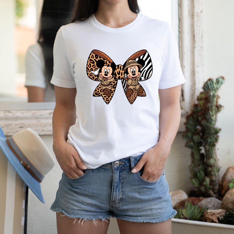 May include: White t-shirt with a large bow design featuring Mickey and Minnie Mouse in safari attire. The bow combines leopard print and zebra stripes. The shirt is worn with denim shorts.