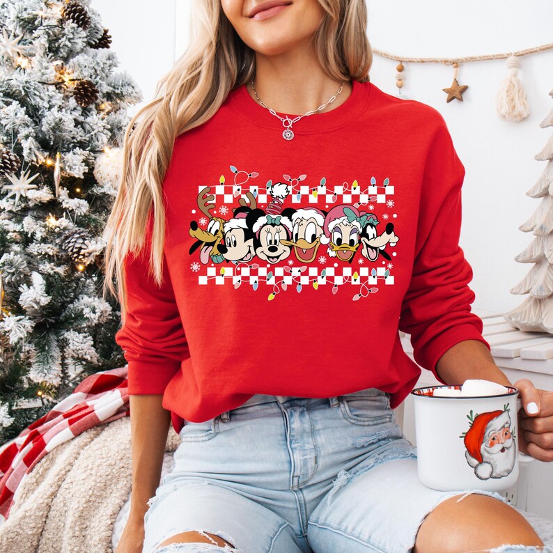 May include: Red Christmas sweater with a checkered pattern and a row of Disney characters including Mickey Mouse, Minnie Mouse, Donald Duck, Goofy, and Pluto. The characters are wearing Santa hats and there are Christmas lights above them.