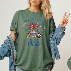 Pode incluir: Camiseta verde musgo com um desenho que apresenta orelhas do Mickey Mouse, um la&ccedil;o vermelho e o texto "Happy 4th of July". O desenho inclui personagens de desenhos animados com roupas patri&oacute;ticas. O nome "Olivia" est&aacute; impresso abaixo.