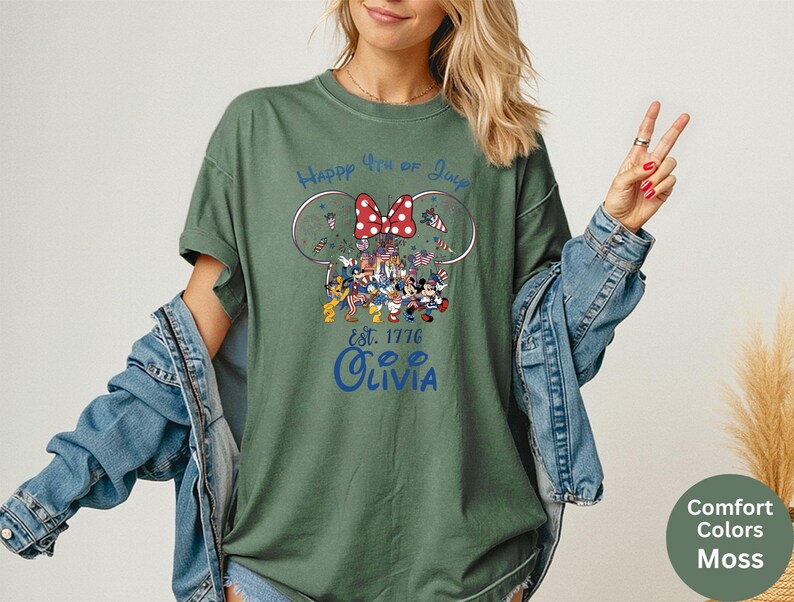 Pode incluir: Camiseta verde musgo com um desenho que apresenta orelhas do Mickey Mouse, um la&ccedil;o vermelho e o texto "Happy 4th of July". O desenho inclui personagens de desenhos animados com roupas patri&oacute;ticas. O nome "Olivia" est&aacute; impresso abaixo.