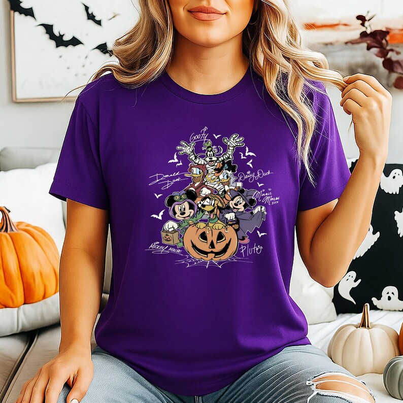 May include: A purple t-shirt featuring a Halloween-themed graphic of Disney characters, including Mickey Mouse, Donald Duck, and Goofy, surrounding a carved pumpkin. The design includes bats and character names in white script.