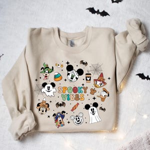 May include: A tan sweatshirt with a Halloween-themed design featuring Mickey Mouse and other Disney characters. The design includes the text "Spooky Vibes" and various Halloween icons such as candy corn, bats, and spiderwebs.