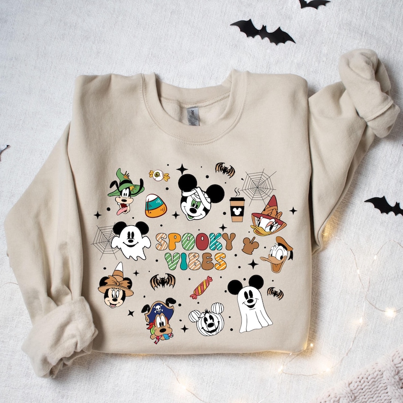 May include: A tan sweatshirt with a Halloween-themed design featuring Mickey Mouse and other Disney characters. The design includes the text "Spooky Vibes" and various Halloween icons such as candy corn, bats, and spiderwebs.