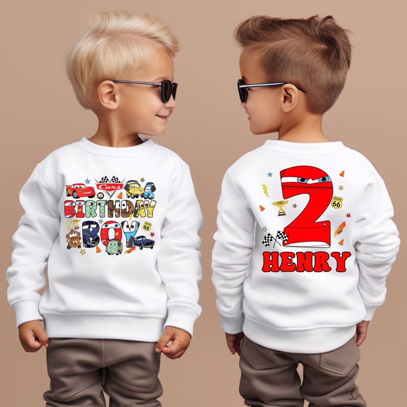 May include: Two white sweatshirts with a Cars theme. The front sweatshirt has a Cars birthday boy design with a red car, a blue car, a yellow car, and a green car. The back sweatshirt has a red number 2 with a car design and the name Henry.