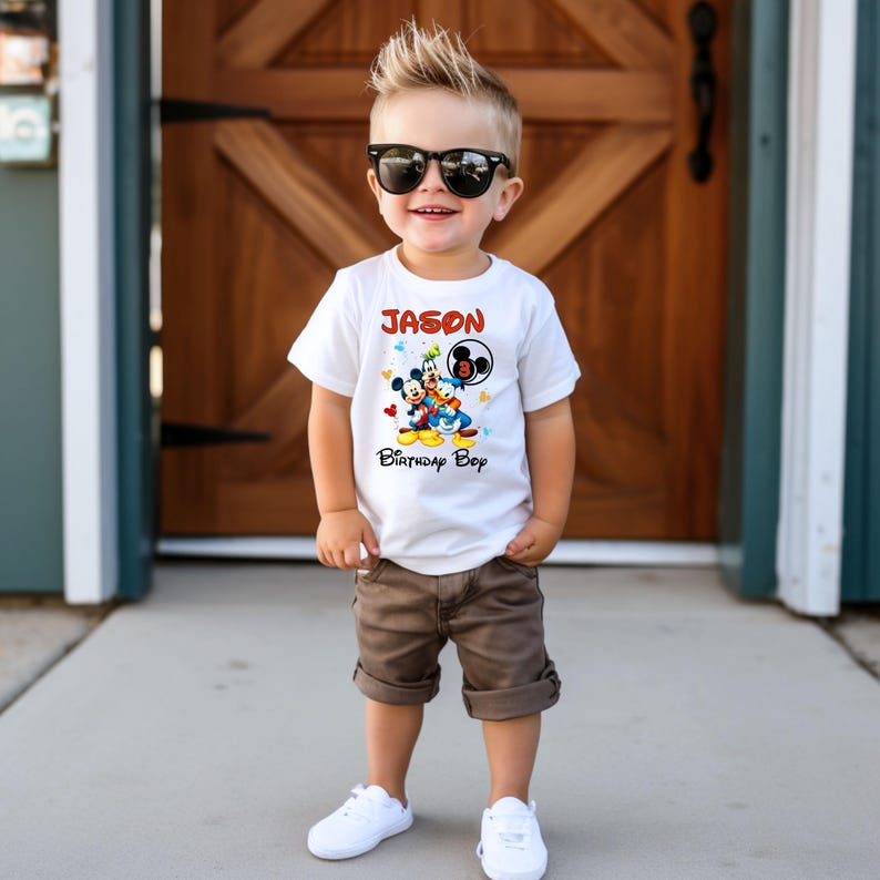 May include: A white t-shirt with a colorful graphic of Mickey Mouse, Donald Duck, and Goofy, with the name "JASON" and "Birthday Boy" printed on it. The child is wearing brown shorts and white sneakers.