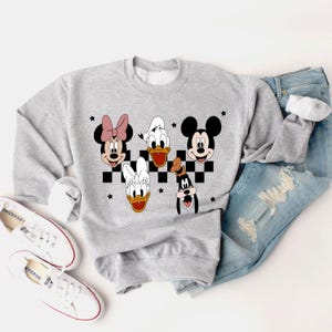 Puede incluir: Una sudadera gris con un patr&oacute;n a cuadros y las caras de Mickey Mouse, Minnie Mouse, Donald Duck, Daisy Duck y Goofy. Los personajes est&aacute;n en blanco y negro con acentos rojos.