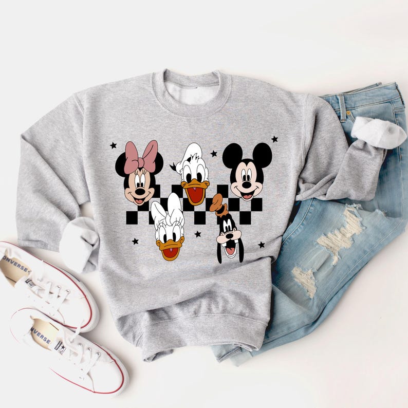 Puede incluir: Una sudadera gris con un patr&oacute;n a cuadros y las caras de Mickey Mouse, Minnie Mouse, Donald Duck, Daisy Duck y Goofy. Los personajes est&aacute;n en blanco y negro con acentos rojos.