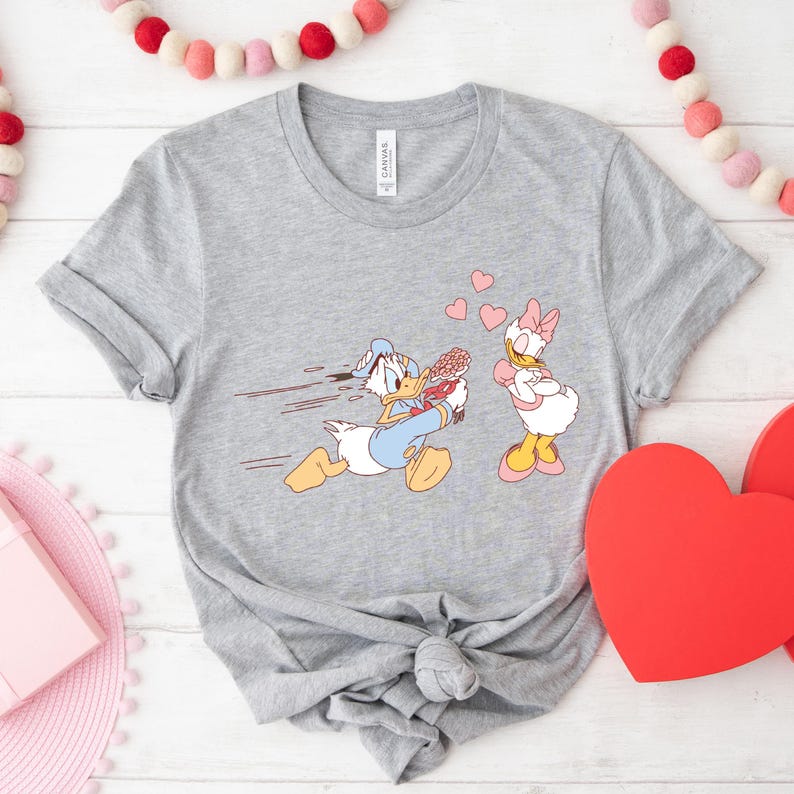Op de afbeelding: Een grijs T-shirt met een cartoon afbeelding van Donald Duck die rent met een bos bloemen naar Daisy Duck, die met gekruiste armen staat en een roze strik in haar haar heeft. Er zweven roze hartjes boven hen.
