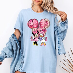 May include: Light blue t-shirt featuring Minnie Mouse and Daisy Duck. The graphic includes a pink bow, castle, and hearts. Minnie Mouse wears a pink polka dot dress, and Daisy Duck wears a purple top. The shirt has short sleeves.