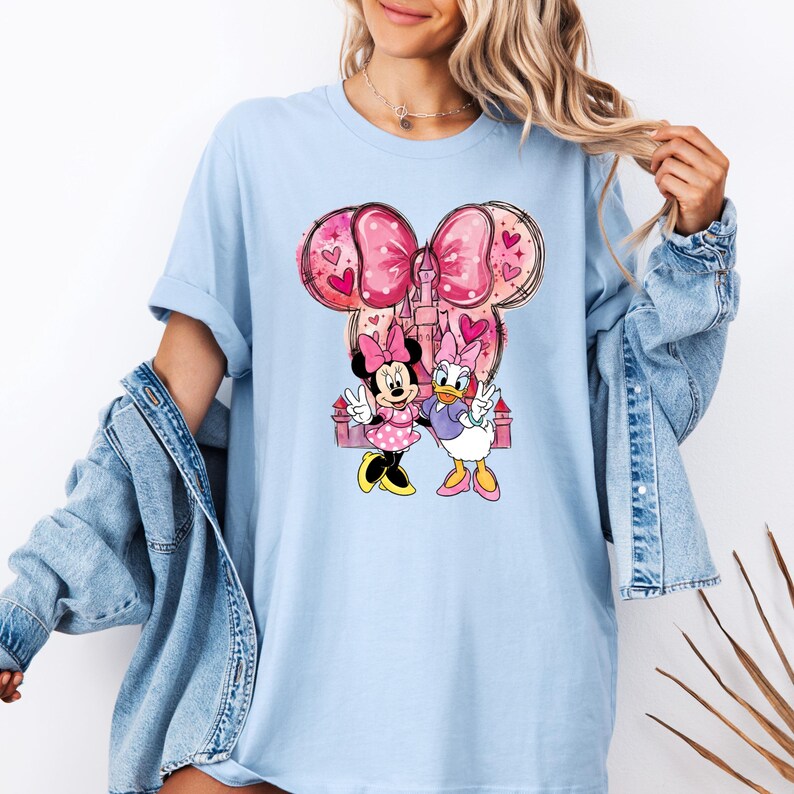 May include: Light blue t-shirt featuring Minnie Mouse and Daisy Duck. The graphic includes a pink bow, castle, and hearts. Minnie Mouse wears a pink polka dot dress, and Daisy Duck wears a purple top. The shirt has short sleeves.