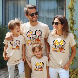Puede incluir: Una familia de cuatro personas con camisetas beige a juego. Cada camiseta presenta un dise&ntilde;o inspirado en Mickey Mouse con un castillo, copos de nieve y la palabra "THANKFUL" en naranja y amarillo. Ideal para una reuni&oacute;n familiar.