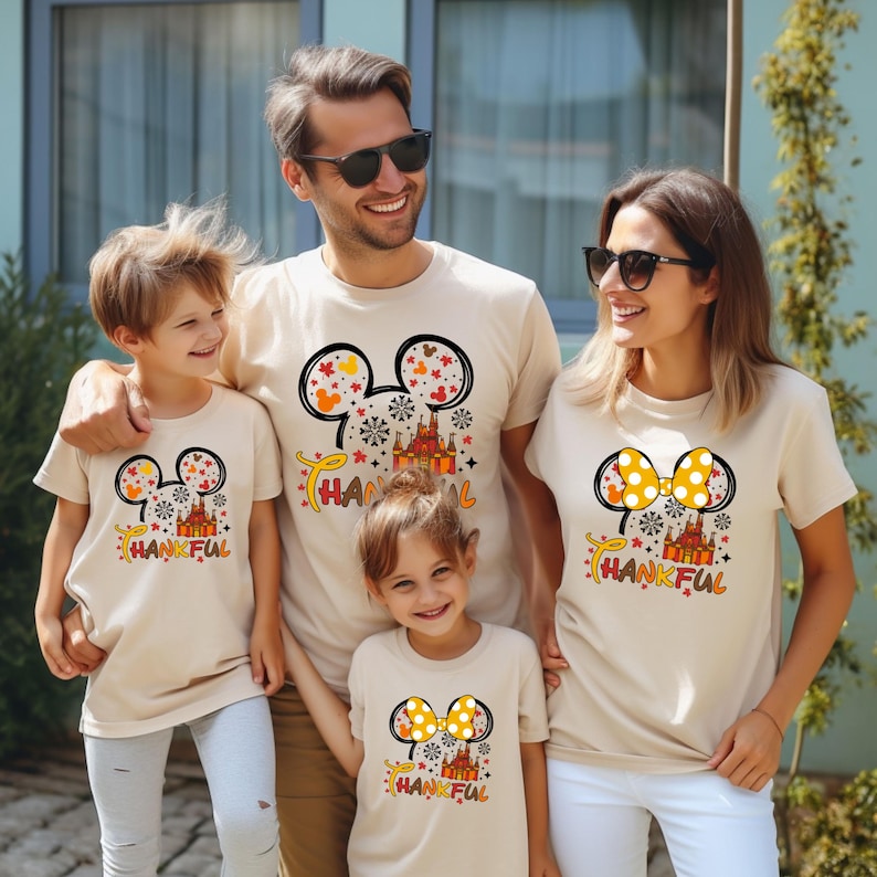 Puede incluir: Una familia de cuatro personas con camisetas beige a juego. Cada camiseta presenta un dise&ntilde;o inspirado en Mickey Mouse con un castillo, copos de nieve y la palabra "THANKFUL" en naranja y amarillo. Ideal para una reuni&oacute;n familiar.