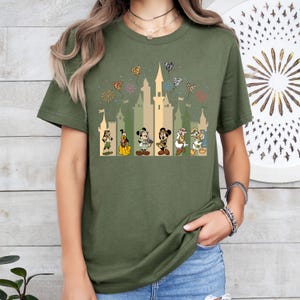 May include: Green t-shirt with a graphic of Mickey Mouse, Minnie Mouse, Donald Duck, Goofy, and Pluto in front of a castle with fireworks and hearts. The characters are dressed in safari outfits.