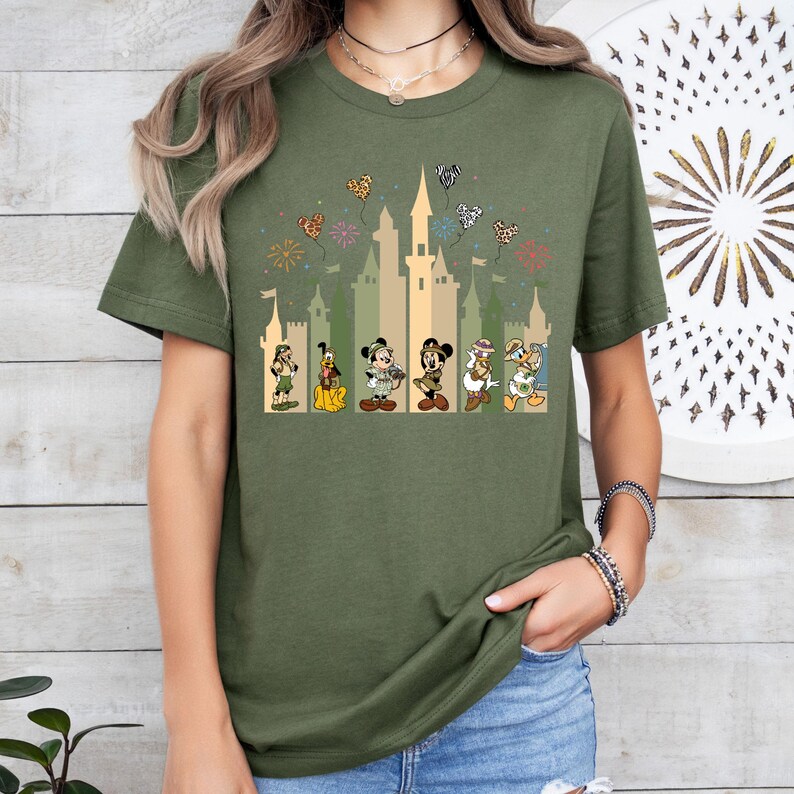 May include: Green t-shirt with a graphic of Mickey Mouse, Minnie Mouse, Donald Duck, Goofy, and Pluto in front of a castle with fireworks and hearts. The characters are dressed in safari outfits.