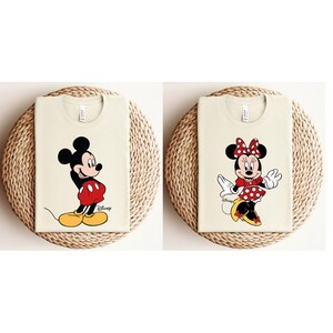 K&ouml;nnte beinhalten: Zwei cremefarbene T-Shirts mit einem Mickey Mouse und Minnie Mouse Grafikdruck. Das Mickey Mouse Shirt hat den Text "Disney" aufgedruckt.