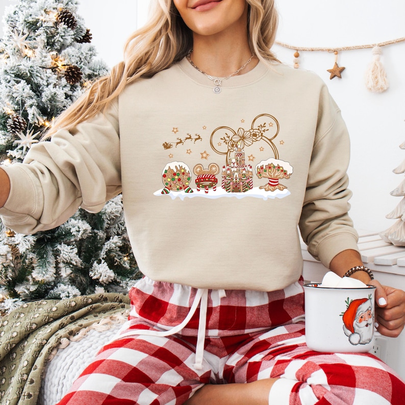 May include: Beige crewneck sweatshirt with a Christmas-themed graphic. The graphic features a Mickey Mouse head made of gingerbread, surrounded by other gingerbread elements, including a mouse, a house, and a tree. The sweatshirt is paired with red and white plaid pajama pants.