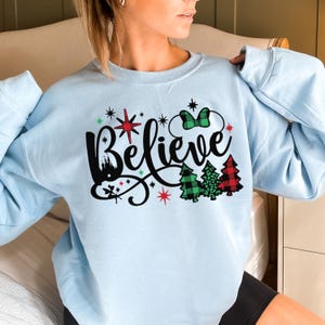 以下が含まれることがあります： 薄い青色のスウェットシャツに、黒と白のグラフィックで「Believe」と書かれており、赤い星、緑と赤の格子縞の木、黒と緑のミッキーマウスのボウが描かれています。