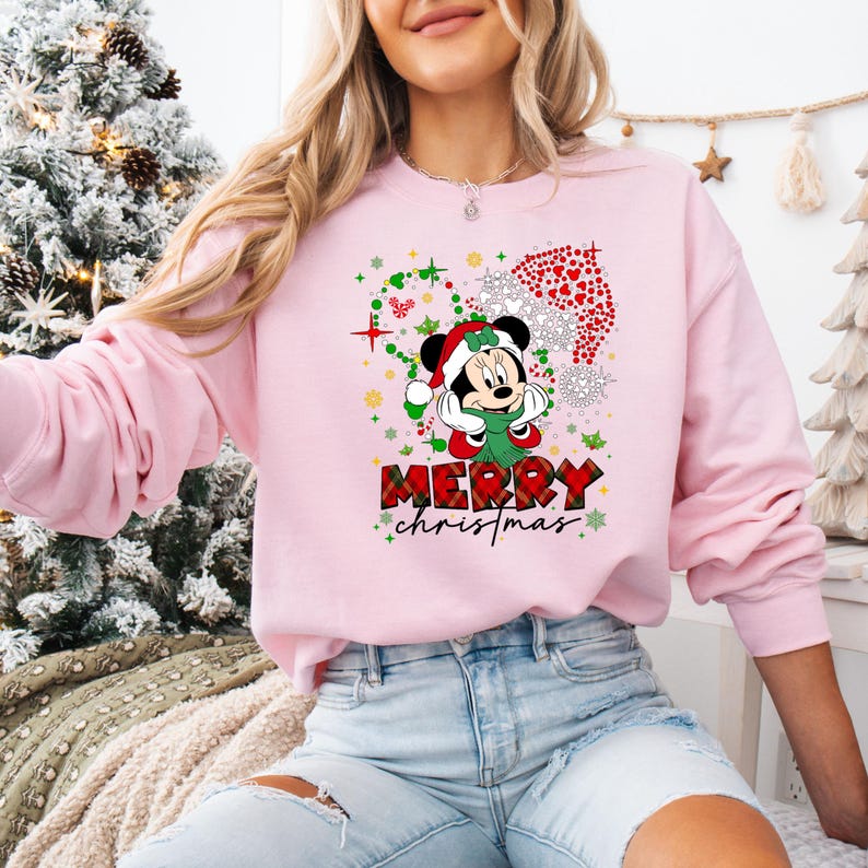 May include: A light pink sweatshirt featuring a graphic of Minnie Mouse in a Santa hat and scarf, surrounded by Christmas decorations. The words "MERRY CHRISTMAS" are printed in a plaid font.