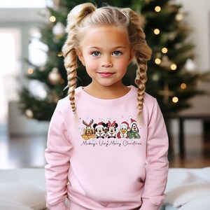 Puede incluir: Sudadera rosa claro con un dise&ntilde;o festivo de personajes de Disney con gorros de Pap&aacute; Noel. El texto "Mickey's Very Merry Christmas" est&aacute; impreso debajo. Llevada por una ni&ntilde;a peque&ntilde;a con trenzas rubias.