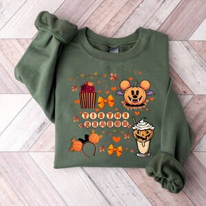 Puede incluir: Sudadera verde con un dise&ntilde;o de Halloween. El dise&ntilde;o incluye un Mickey Mouse con forma de calabaza, una magdalena, una bebida y el texto "Tis the Season". Los lazos y hojas naranjas completan el look festivo.