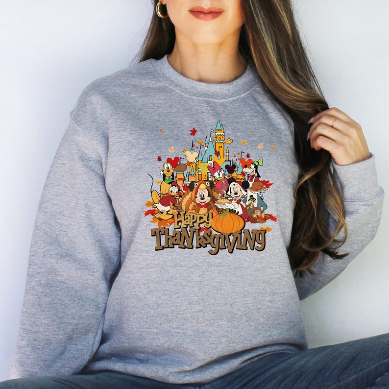 May include: A gray sweatshirt featuring a colorful graphic of Disney characters gathered for Thanksgiving. The design includes a pumpkin, autumn leaves, and the text "Happy Thanksgiving". A castle is in the background.