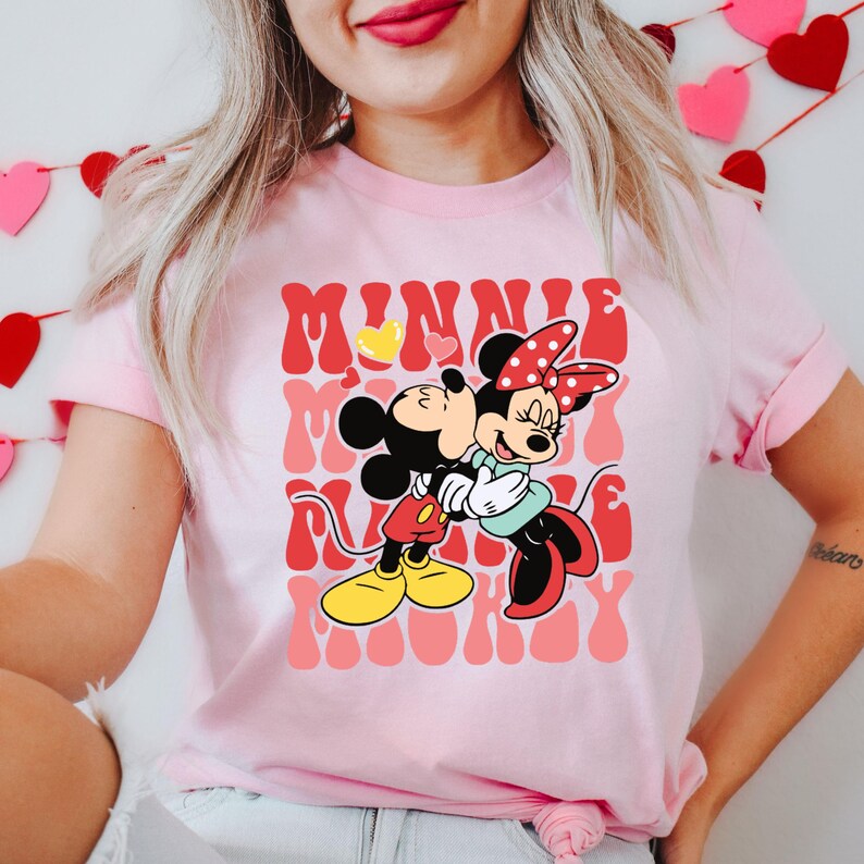 K&ouml;nnte beinhalten: Hellrosa T-Shirt mit einer Grafik von Mickey und Minnie Mouse, die sich umarmen. Das Design enth&auml;lt die W&ouml;rter "MINNIE" und "MICKEY" in einer Retro-Schriftart mit einem Herz-Akzent. Die Charaktere tragen ihre klassischen Outfits.